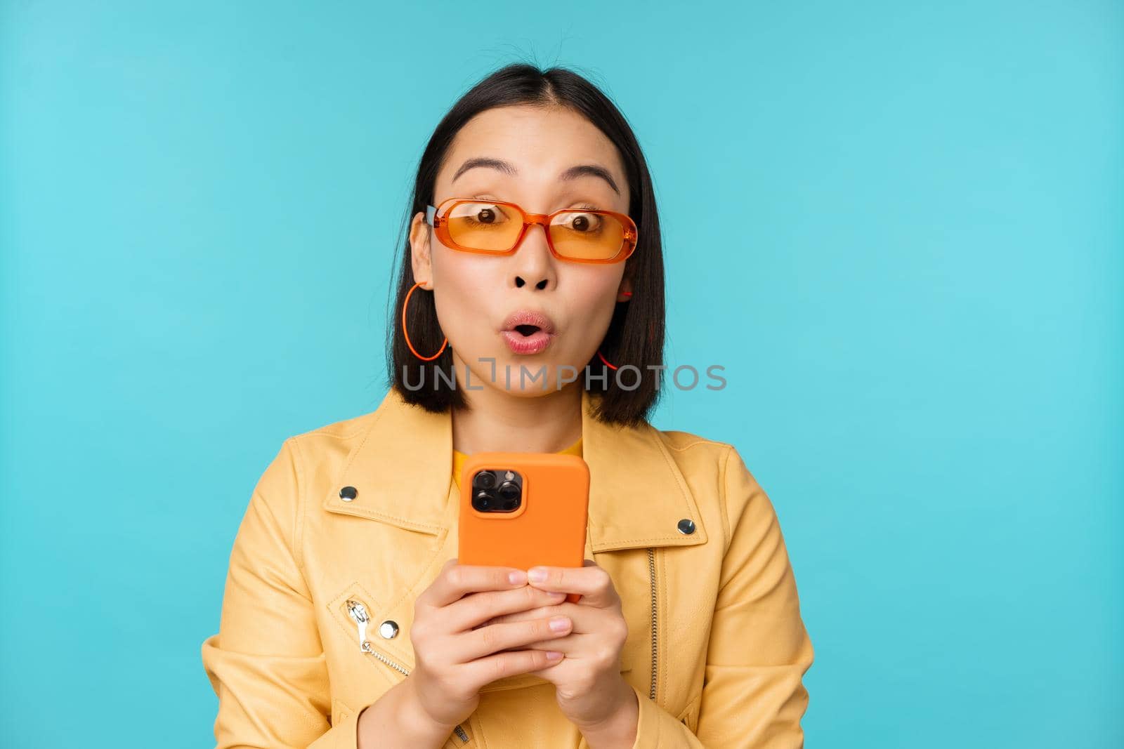 Image of asian girl in sunglasses, looking amazed and impressed, recording video or taking photo on smartphone, standing over blue background.