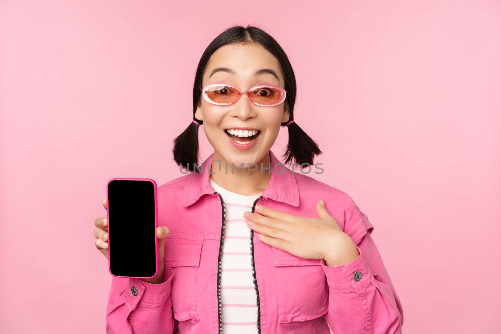 Image of surprised girl showing mobile phone app screen, smartphone display, application interface, standing against pink background by Benzoix