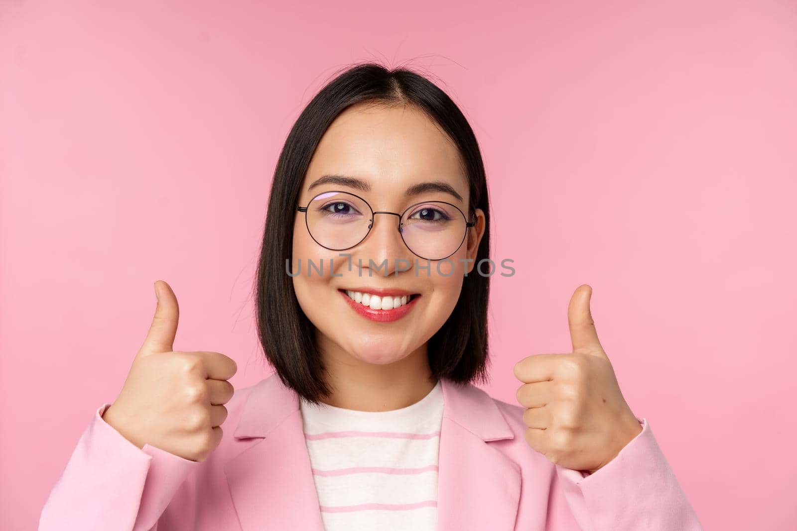 Awesome, congrats. Face of excited asian businesswoman in glasses, smiling pleased, showing thumbs up in approval, standing over pink background by Benzoix