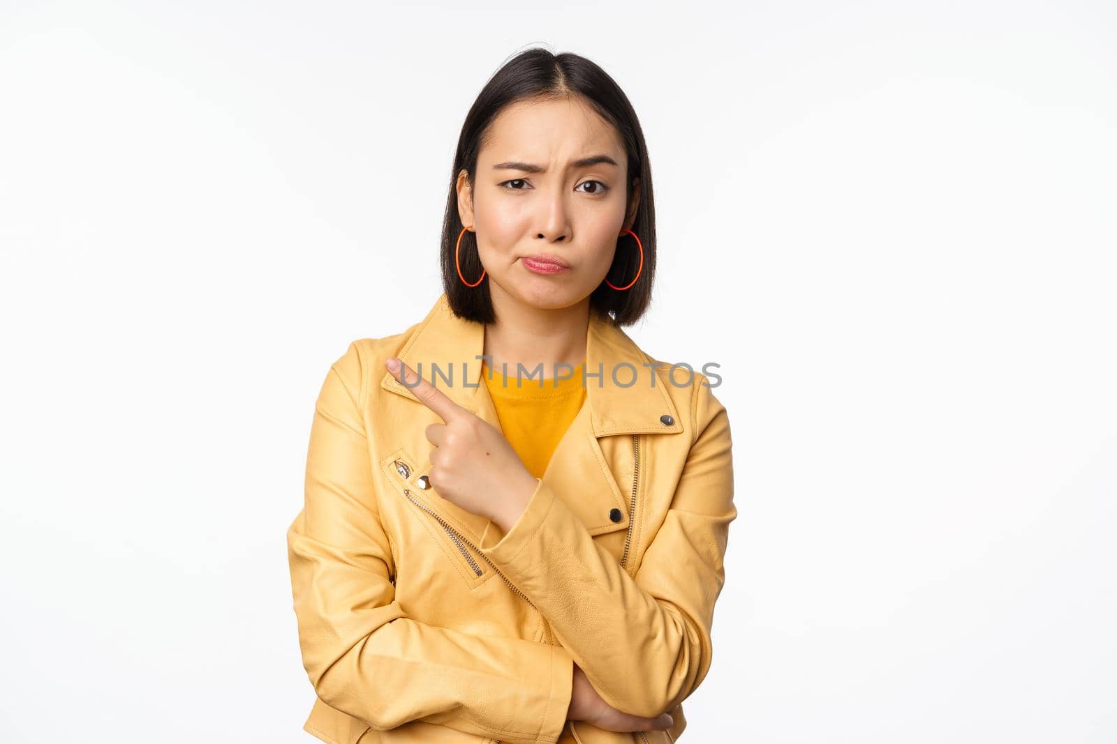 Portrait of stylish asian girl looking with doubt, skeptical face, pointing finger left at logo or advertisement, standing against white background by Benzoix