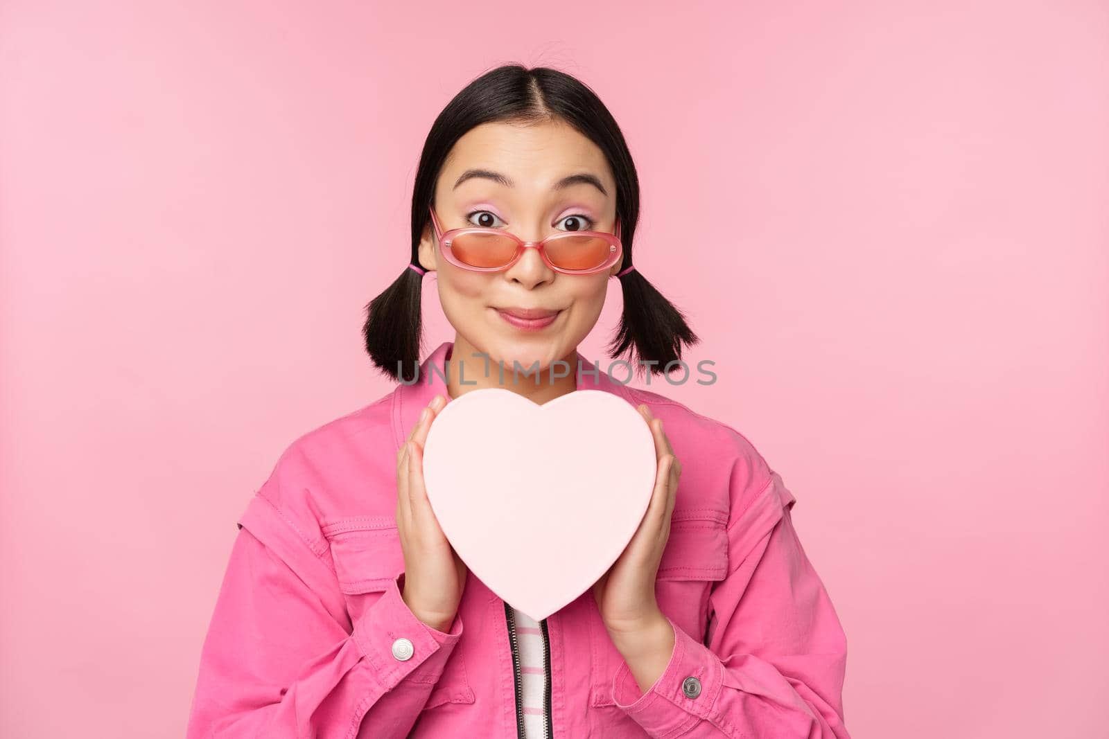 Beautiful asian girl smiling happy, showing heart gift box and looking excited at camera, standing over pink romantic background.