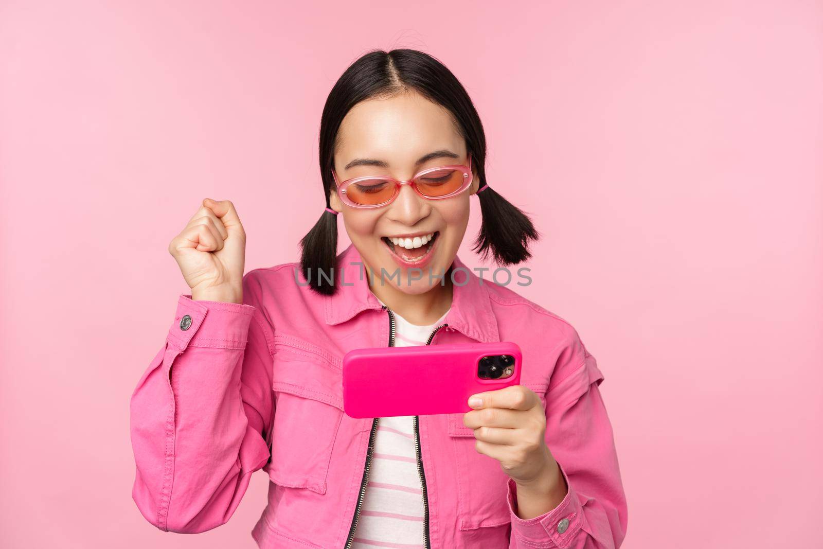 Happy smiling korean girl winning on mobile phone, looking at horizontal smartphone screen and rejoicing, achieve goal, celebrating, standing over pink background.