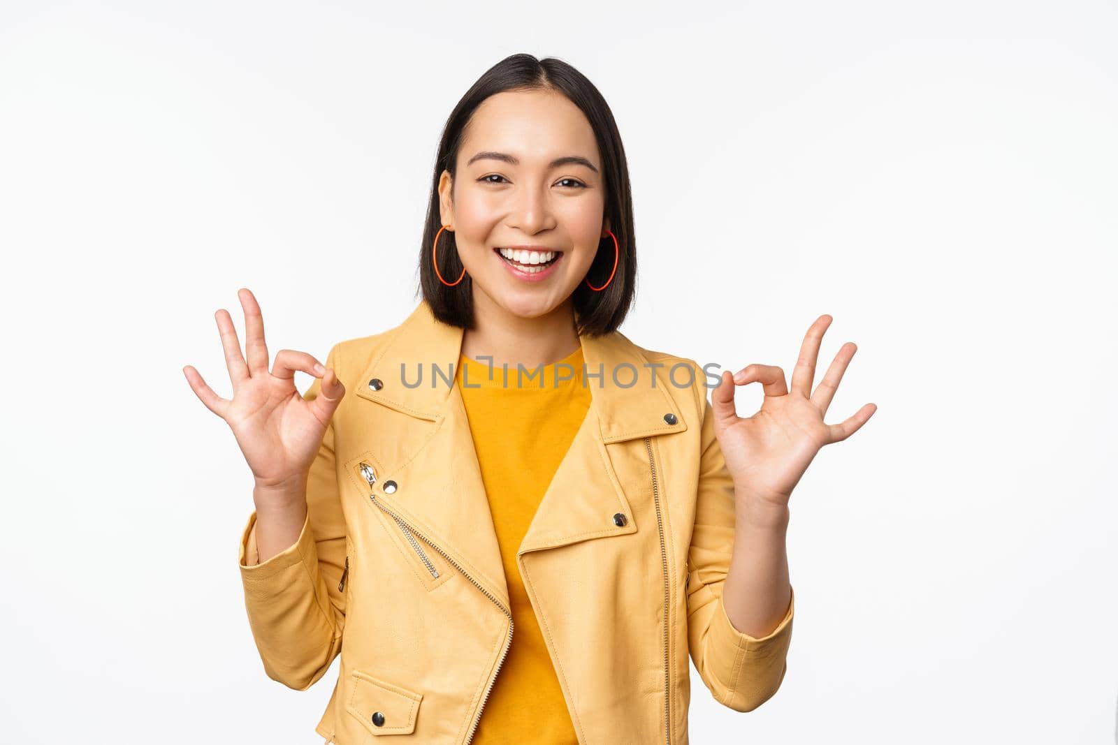 Happy beautiful asian girl shows okay, ok recommending sign, pleased by smth, praising quality, standing over white background. Copy space
