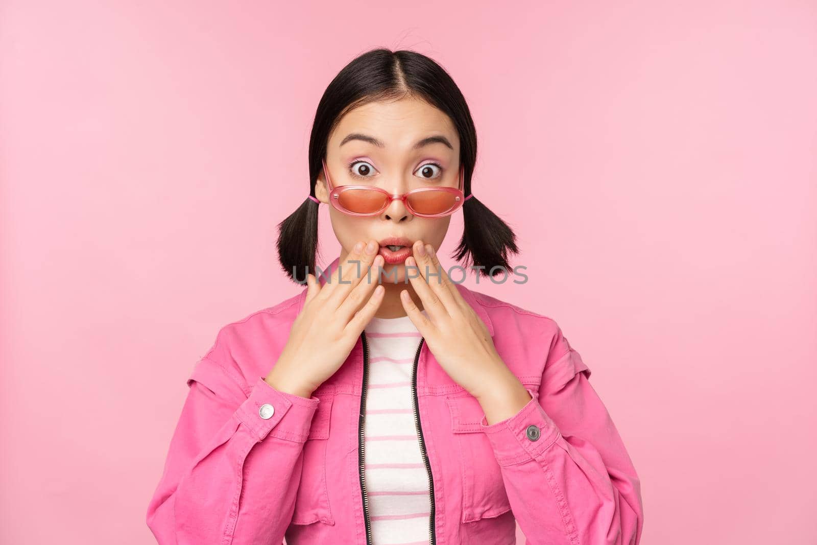 Portrait of young asian woman in stylish sunlgasses, looking surprised, disbelief reaction, standing over pink background by Benzoix