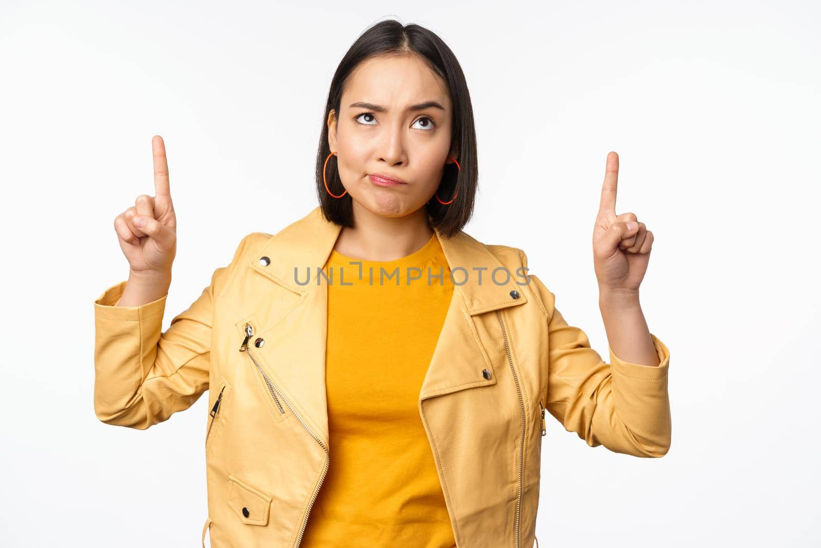 Disappointed korean female model, frowning and grimacing skeptical, pointing up and looking upset at banner on top, standing over white background by Benzoix