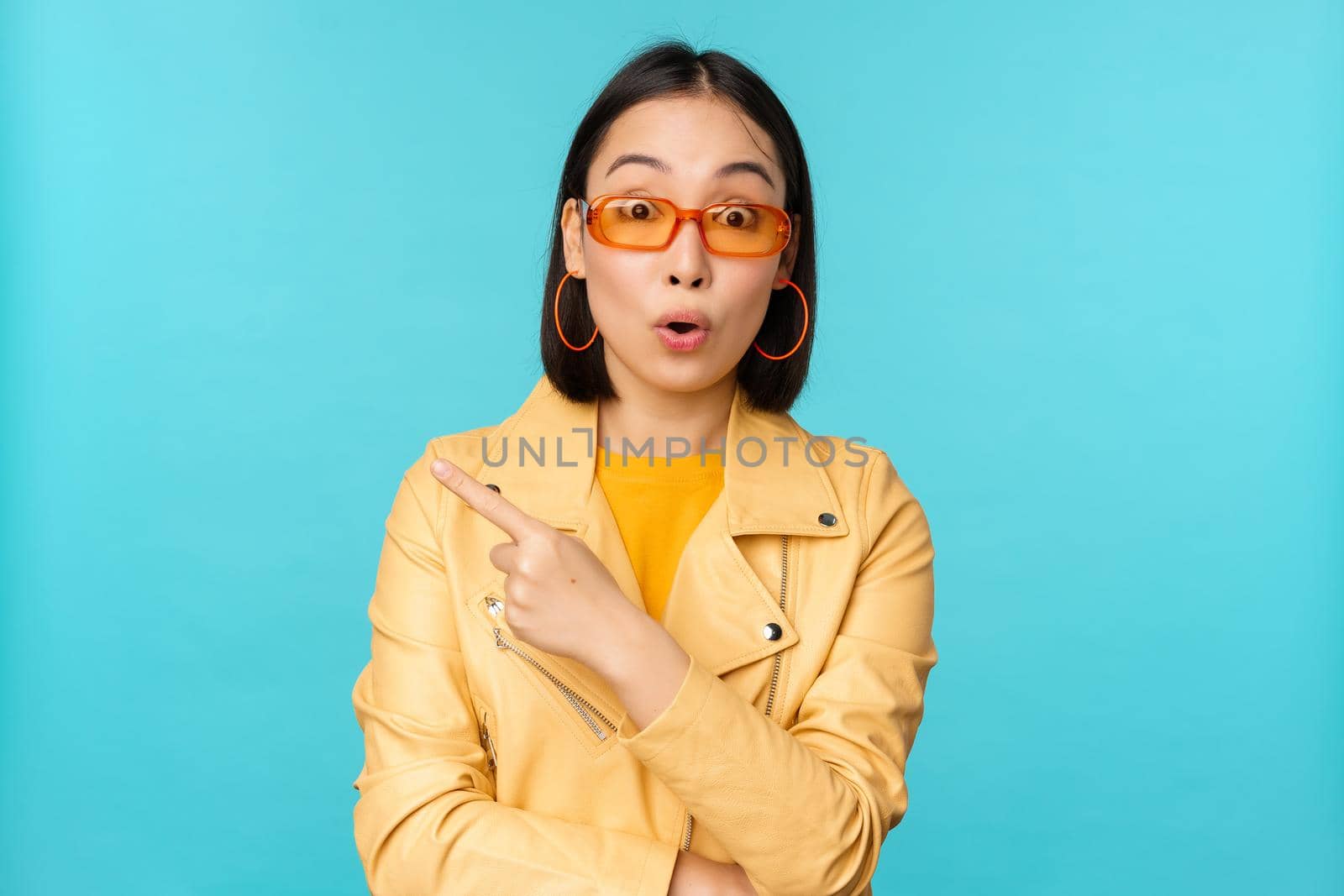 Image of asian woman looks intrigued, asks question about item or store, points finger left with surprised face expression, stands over blue background.