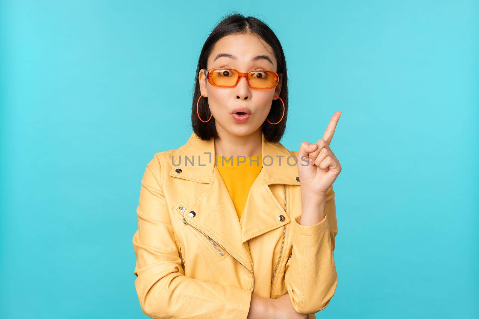 Excited asian girl raising finger, suggesting smth, eureka sign, has plan, standing in sunglasses over blue background by Benzoix