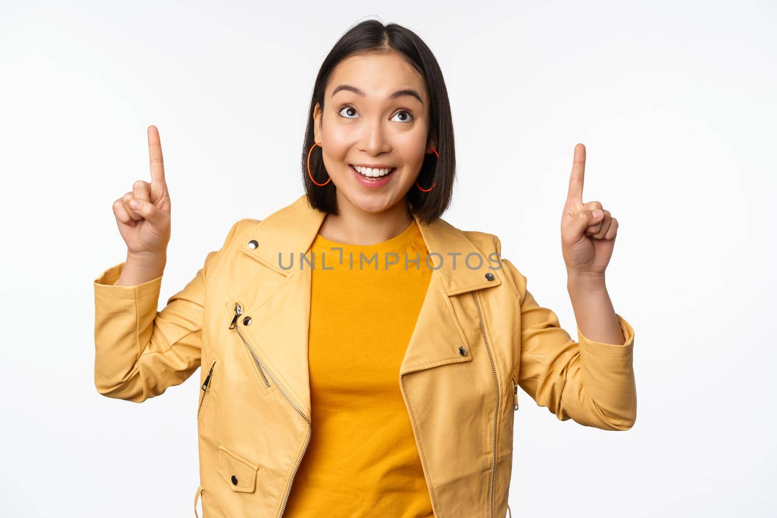 Image of smiling asian brunette woman pointing fingers up, showing advertisement with happy face, posing against white background by Benzoix