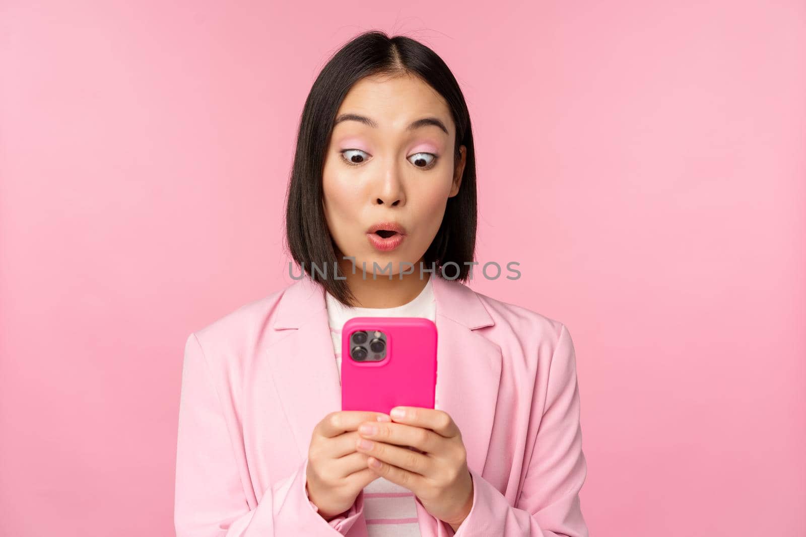 Portrait of asian businesswoman with surprised face, using smartphone app, wearing business suit. Korean girl with mobile phone and excited face expression, pink background.