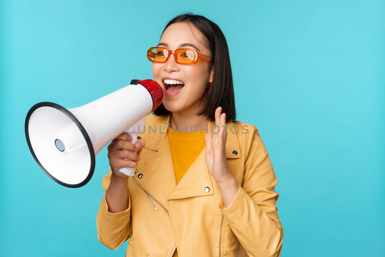 Stylish asian girl making announcement in megaphone, shouting with speakerphone and smiling, inviting people, recruiting, standing over blue background.