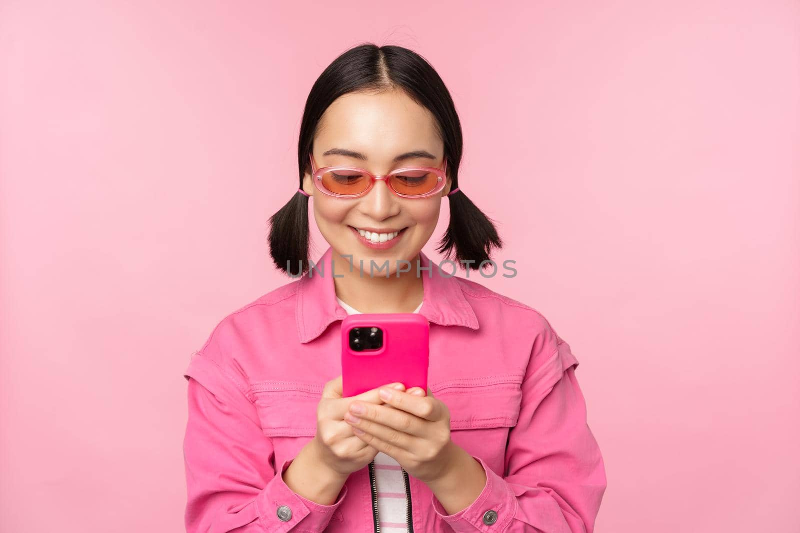 Portrait of asian girl in sunglasses using smartphone. Woman looking at mobile phone, browsing in app, standing over pink background.