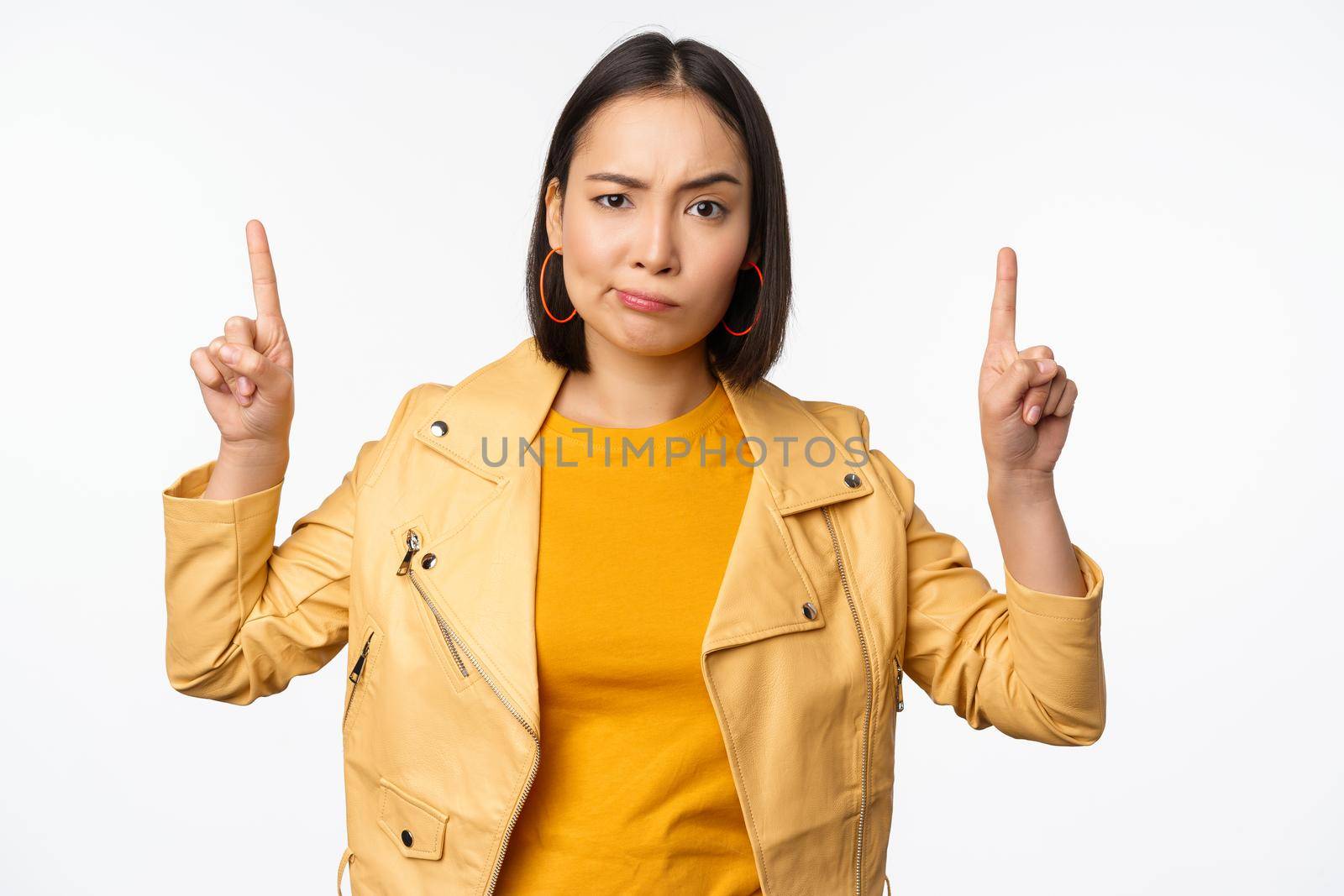 Skeptical asian woman pointing fingers up, frowning and looking with disapproval, complaining at smth, standing doubtful against white background by Benzoix