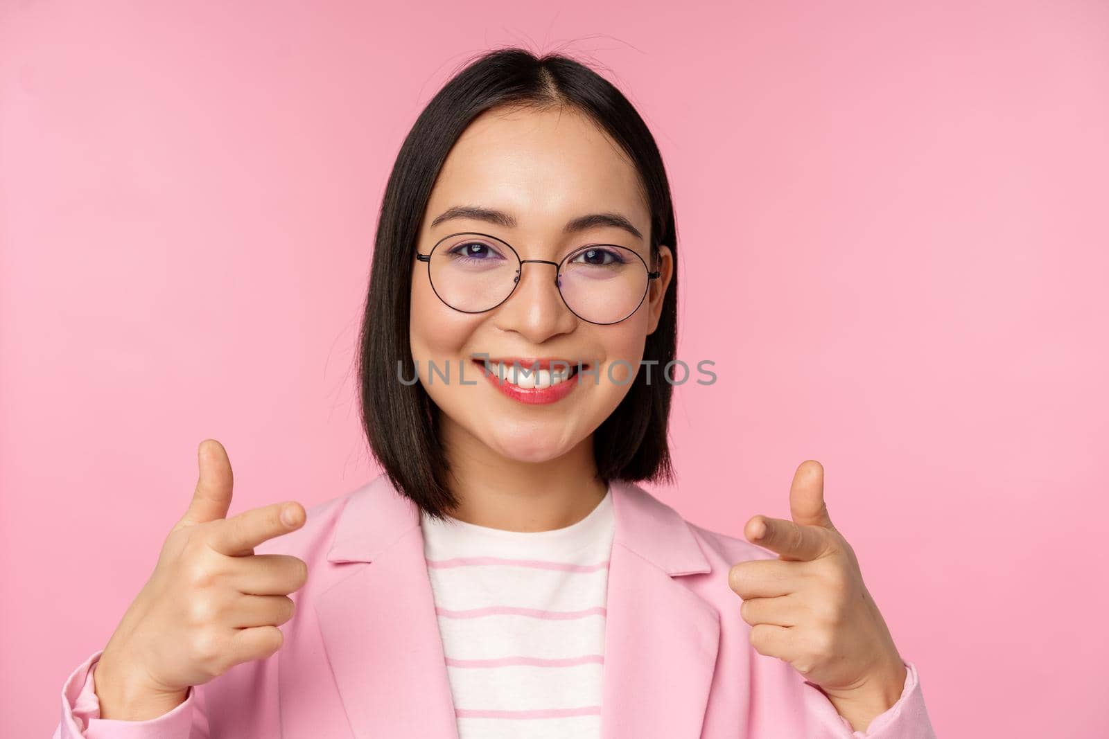 Its you, congratulations. Smiling asian corporate woman, ceo manager in suit and glasses, pointing fingers at camera, recruiting, praise or compliment, standing over pink background by Benzoix