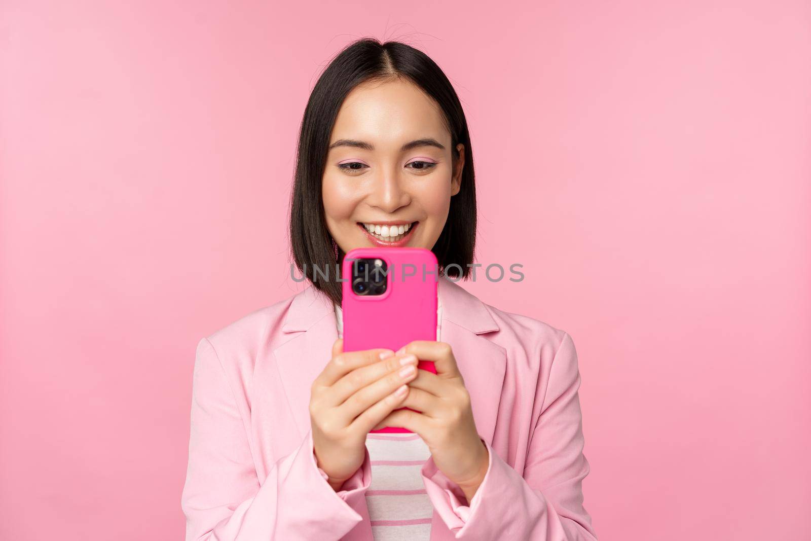 Image of smiling asian corporate woman in suit looking, watching on smartphone app, using mobile phone application, standing over pink background.