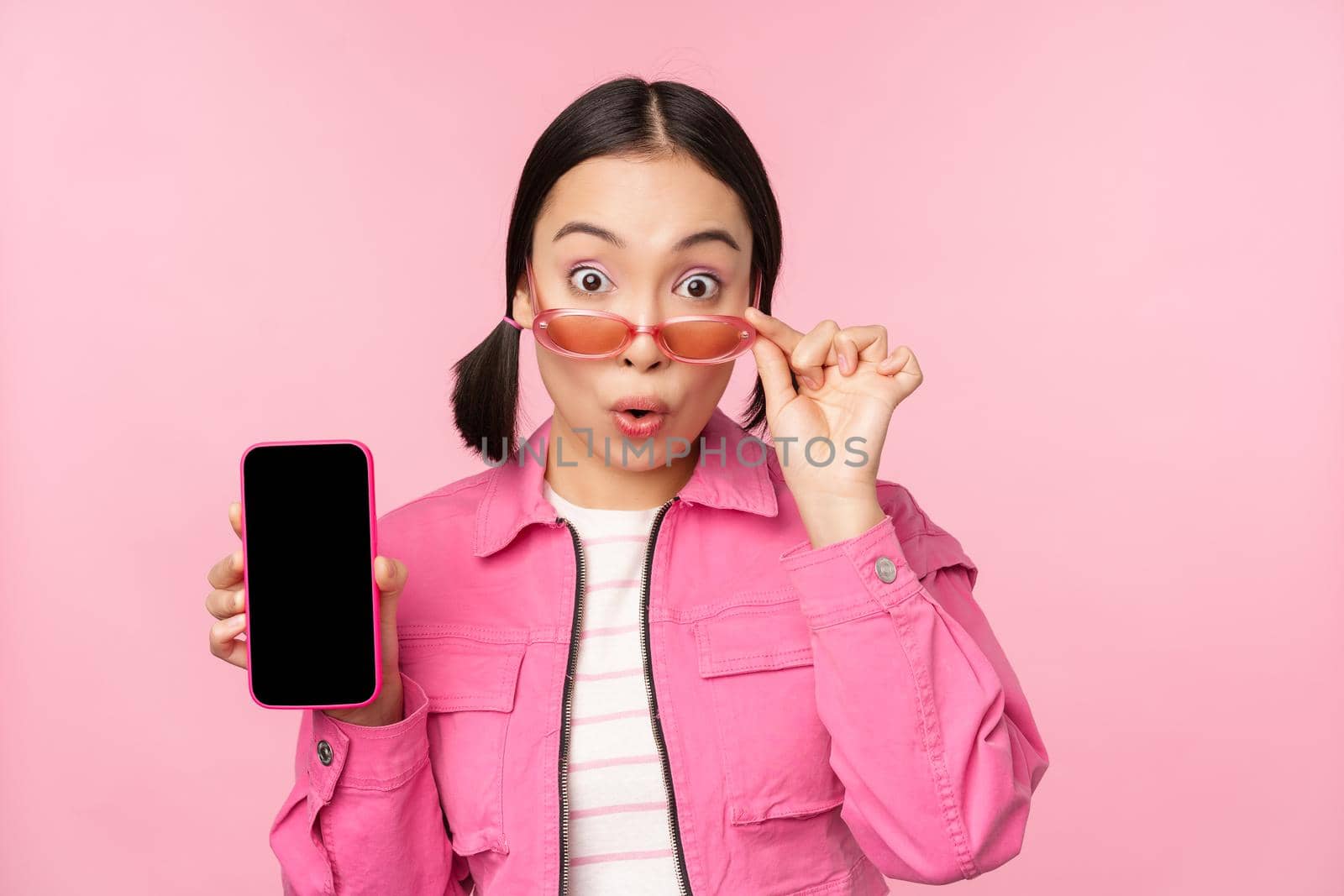 Portrait of asian girl showing mobile phone screen, reacting surprised, standing over pink background by Benzoix