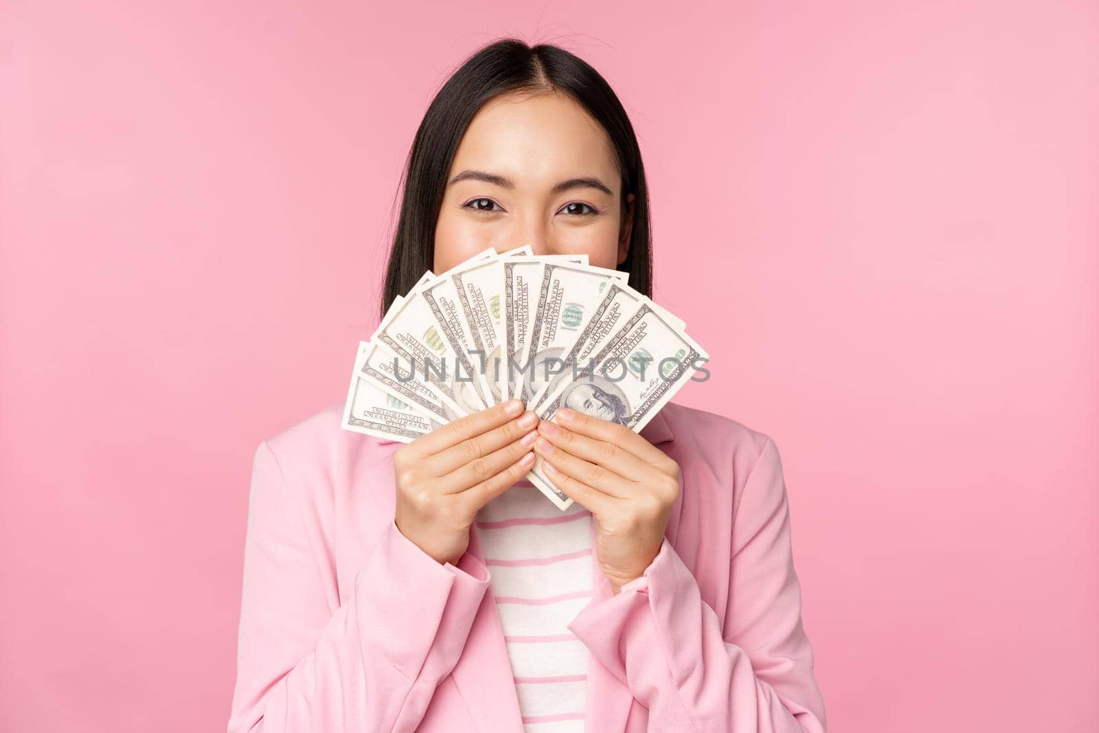 Happy asian lady in suit holding money, dollars with pleased face expression, standing over pink background. Copy space