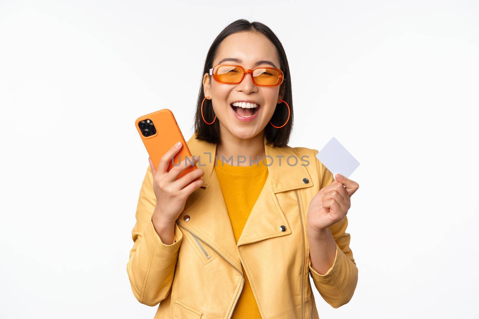 Online shopping and delivery concept. Happy korean girl in stylish clothes, holding credit card and smartphone, laughing and smiling, standing over white background by Benzoix