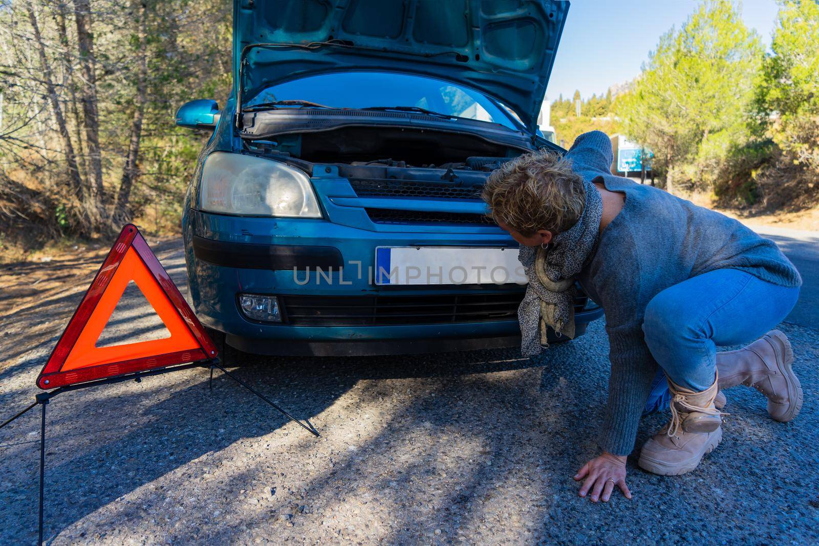Caucasian blonde adult female observing engine failure in her car while traveling. Small blue car with engine failure. Modern and autonomous adult woman trying to fix her car on the road. She has red safety triangle on the ground. Very sunny clear day.