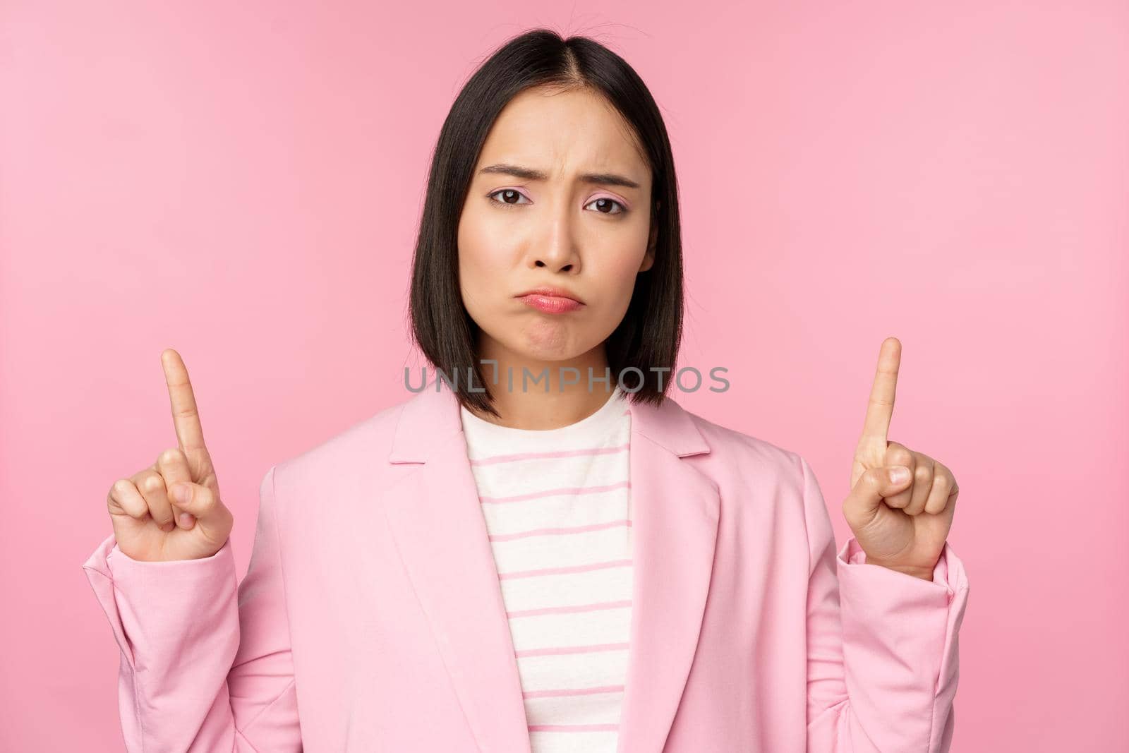 Sad asian business woman, professional corporate manager pointing fingers up, sulking disappointed, standing over pink background.