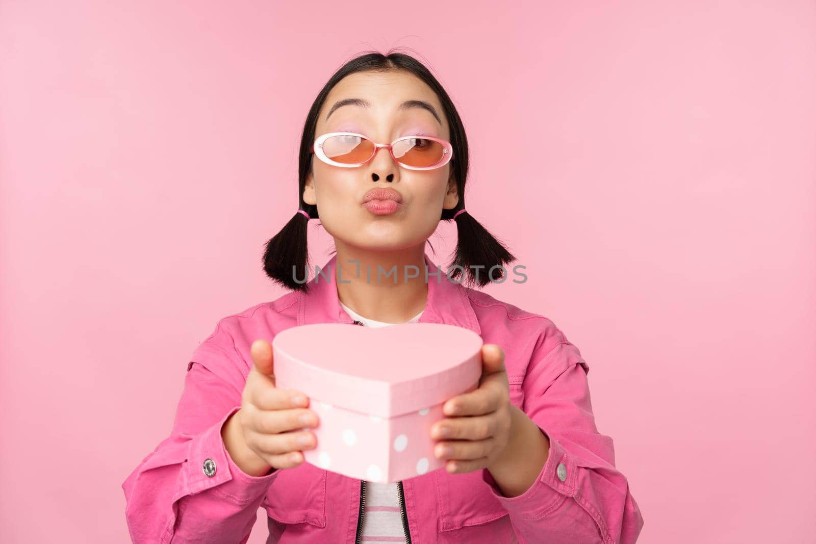 Cute asian girl giving you gift in heart shaped box, kissing and smiling, concept of holiday and celebration, standing over pink background by Benzoix