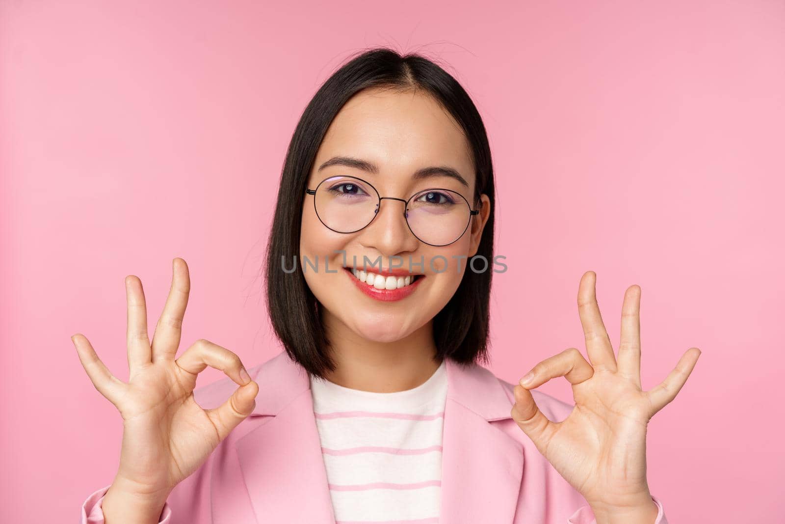 Close up portrait of impressed corporate woman, asian business lady in glasses, showing okay sign, looking amazed at camera, recommending, pink background by Benzoix