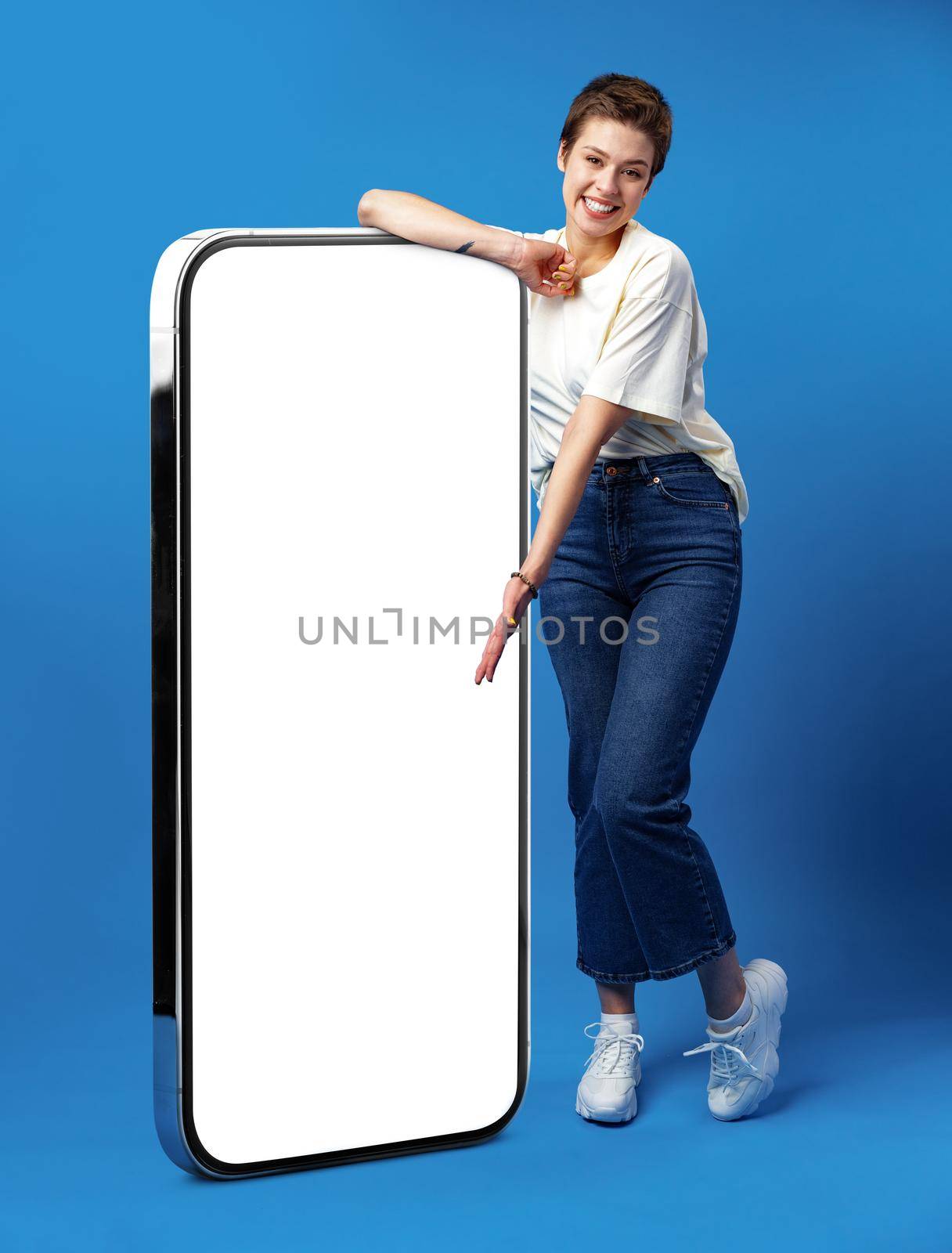 Happy young woman leaning on huge cellphone with empty white screen, for your ad, mock up, blue backgorund