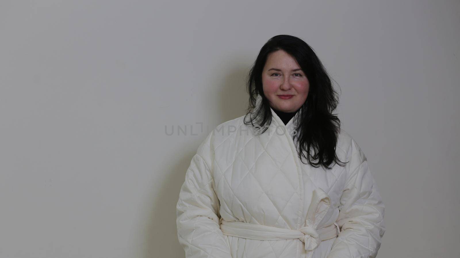 Smiling beautiful dark-haired plus size body positive woman in a white winter coat looking at the camera, against the wall...Brunette against a white wall in a white coat.