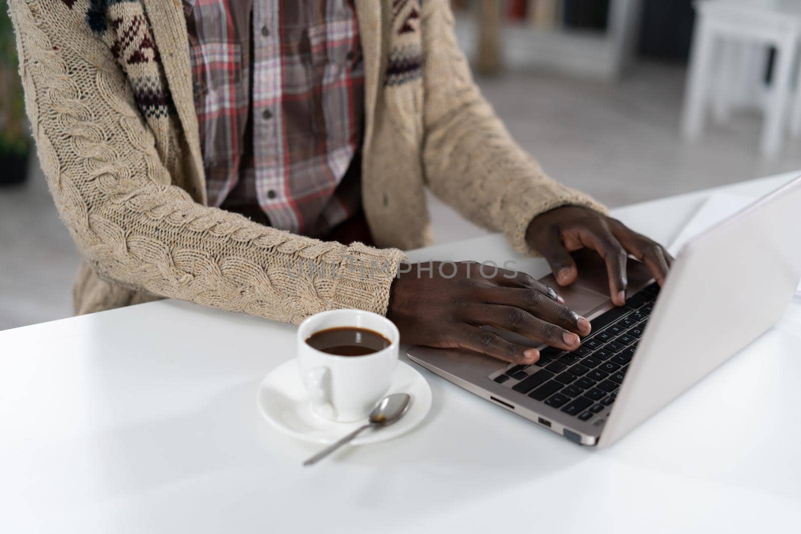 African American man typing on laptop with a cup of coffee next to him wearing beige sweater sitting at the table at the campus cafeteria. Drops of coffee on spoon isolated on white table.