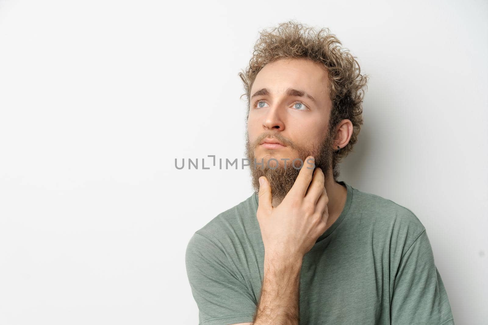 Thoughtful or pensive young handsome bearded wild curly hair man with bright blue eyes isolated on white background. Young thinking man in green t shirt on white. Copy space.