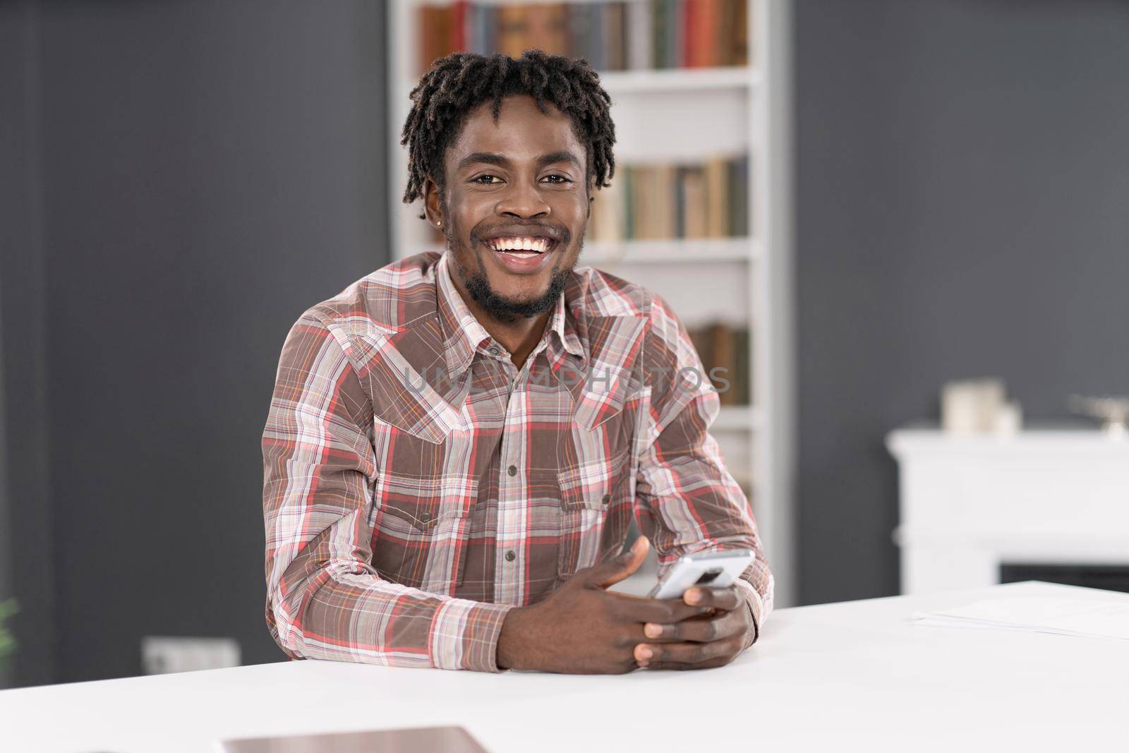 Smiling handsome African American man use smartphone checking social media or updating information. Portrait of young man holding smartphone sitting at the table. Social media concept.