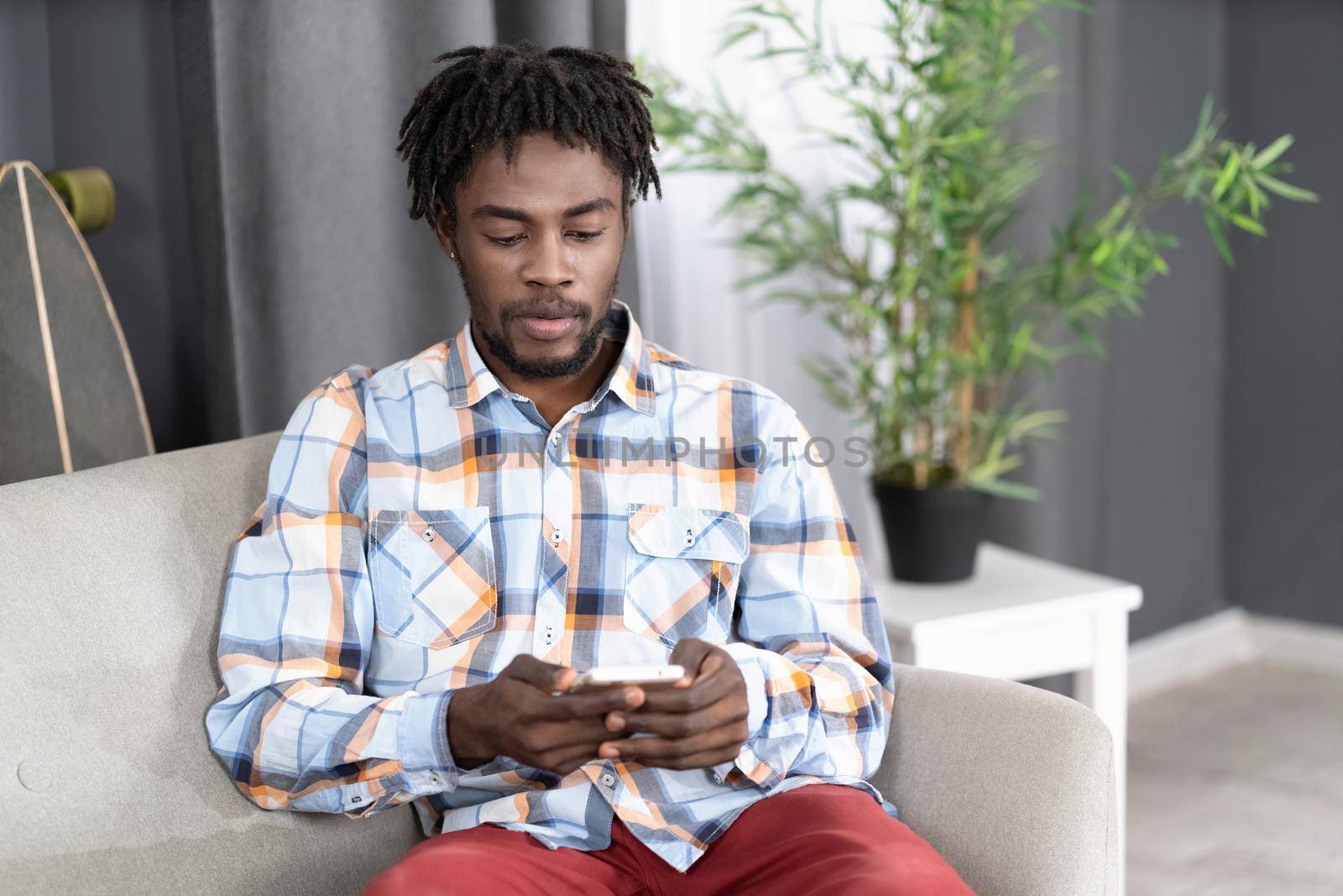 Concentrated or interested African American man use smartphone checking social media or updating information. Portrait of young man holding smartphone sitting on the sofa. Social media concept.