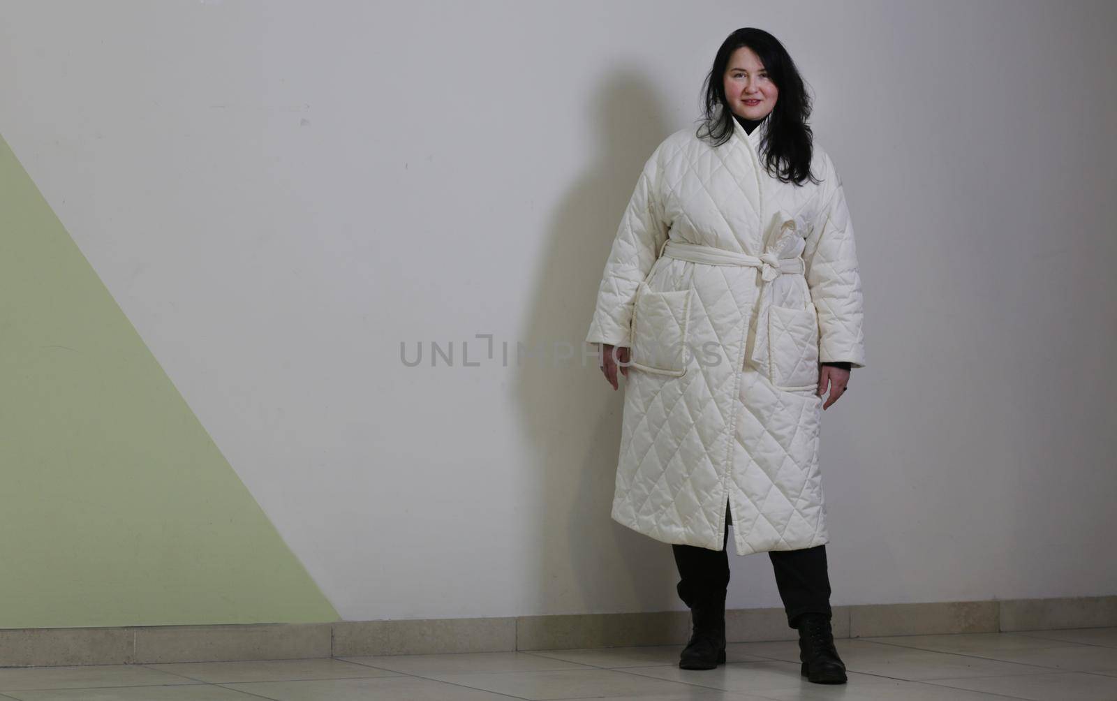 An attractive woman plus size in a white winter coat against a light wall poses and looks at the camera.