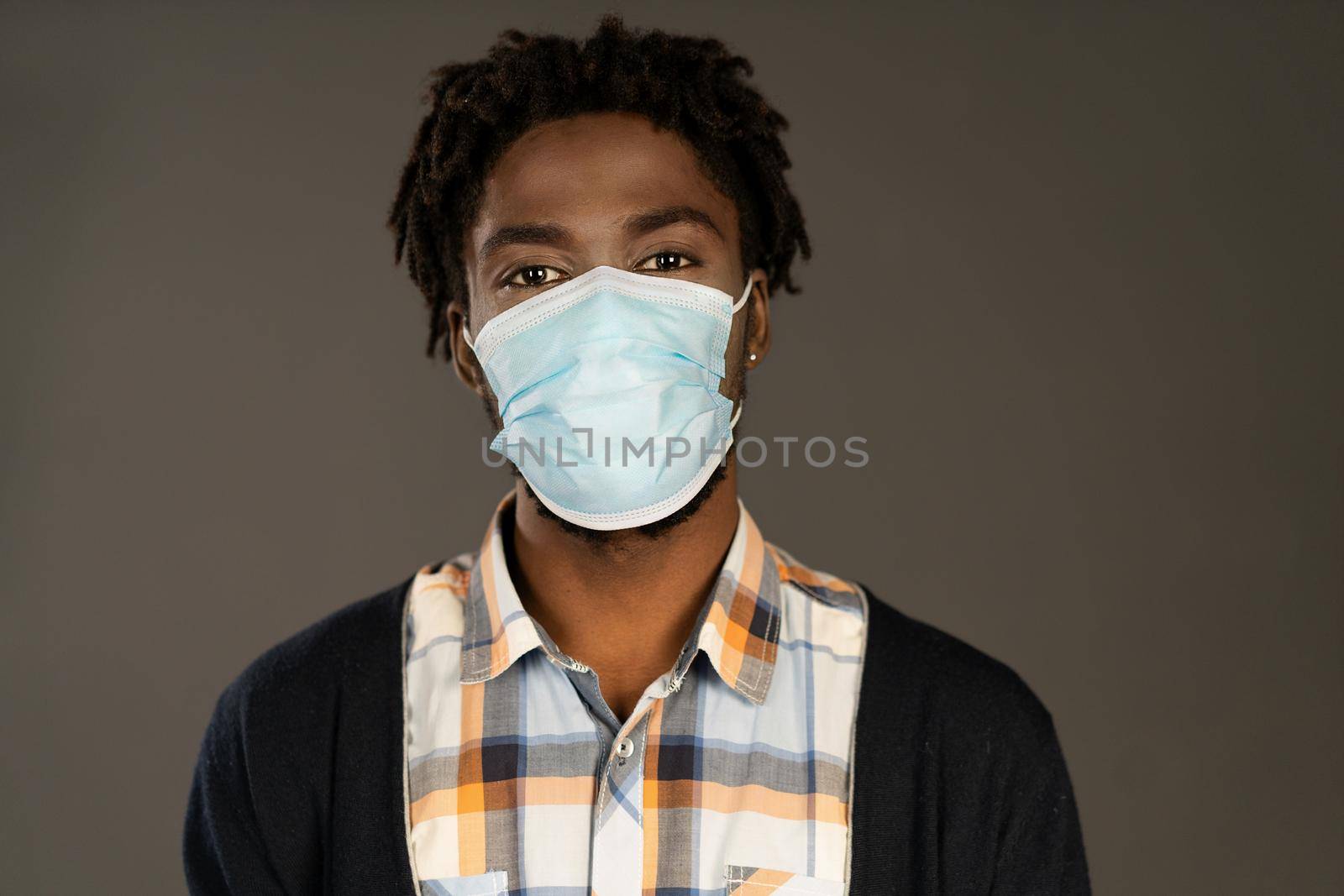 Protective mask on face of African-american man looking at camera. Happy or tired young man wearing medical mask. African man with medical mask oh his face. Isolated on grey background.