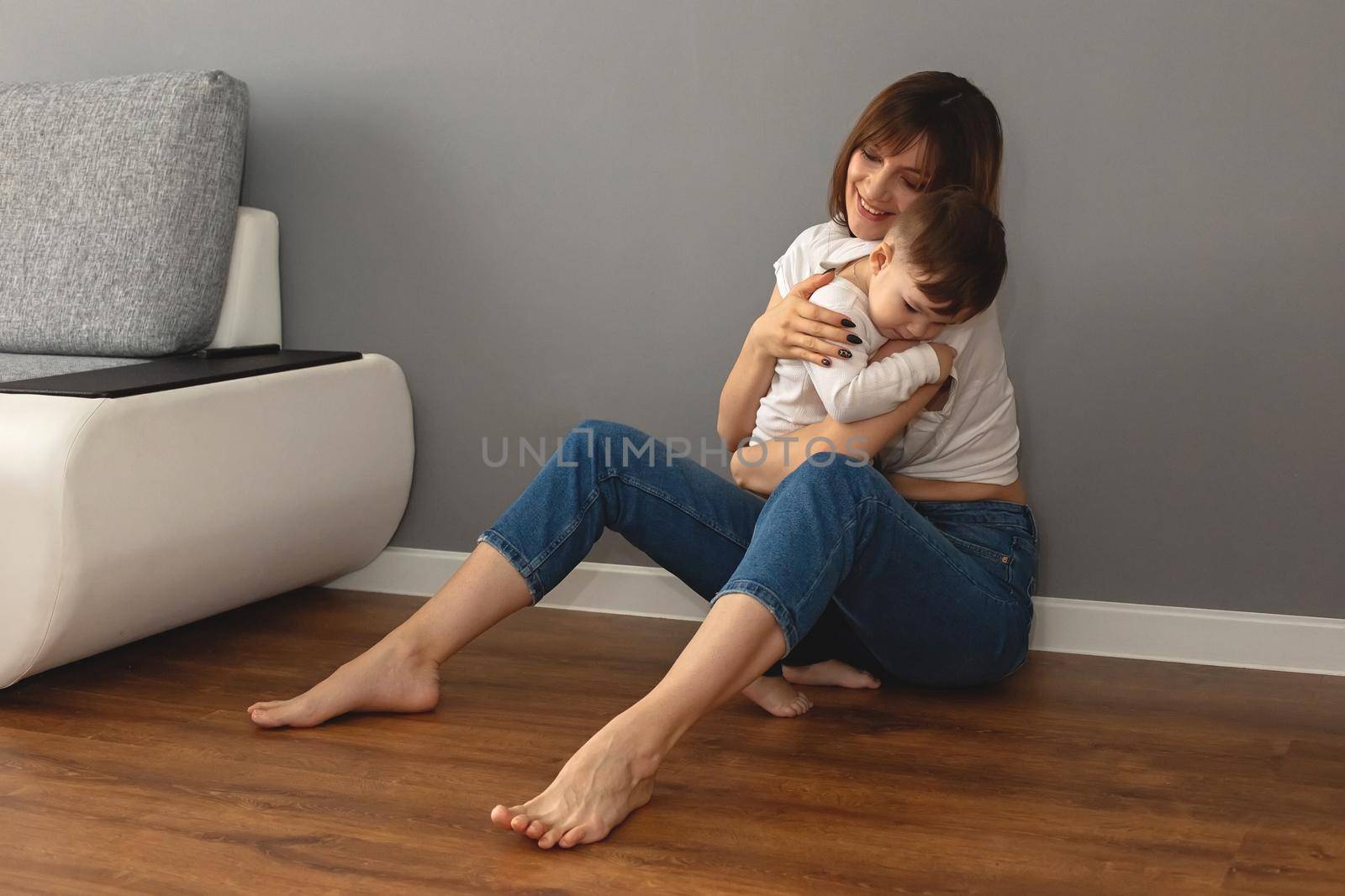 Mom and baby are sitting on the floor in the room, near the gray wall, gently hugging each other. Copy space