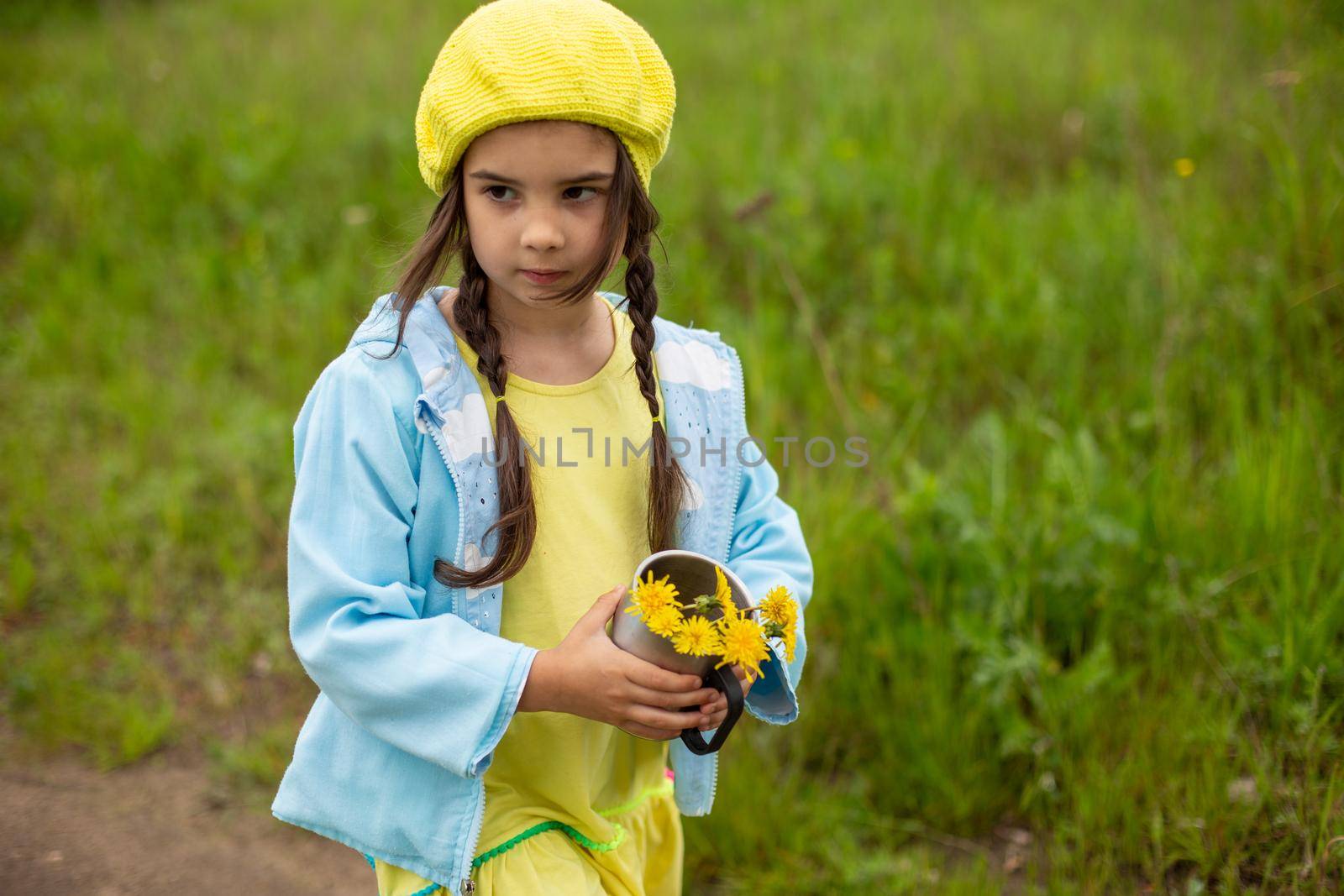 A little girl in a yellow knitted beret and a blue jacket walks along a path near a green lawn with a bouquet of dandelions