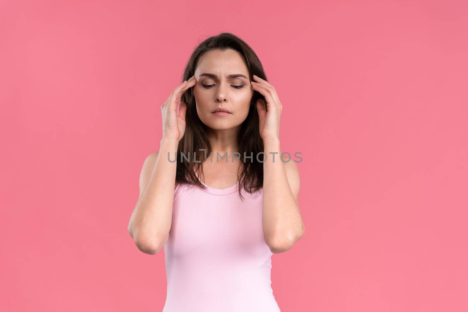 Suffering of headache or migraine young woman in sleeveless pink t-shirt holding her head with eyes shut isolated on pink background. Stressed employee woman in stripe suit. With copy space.