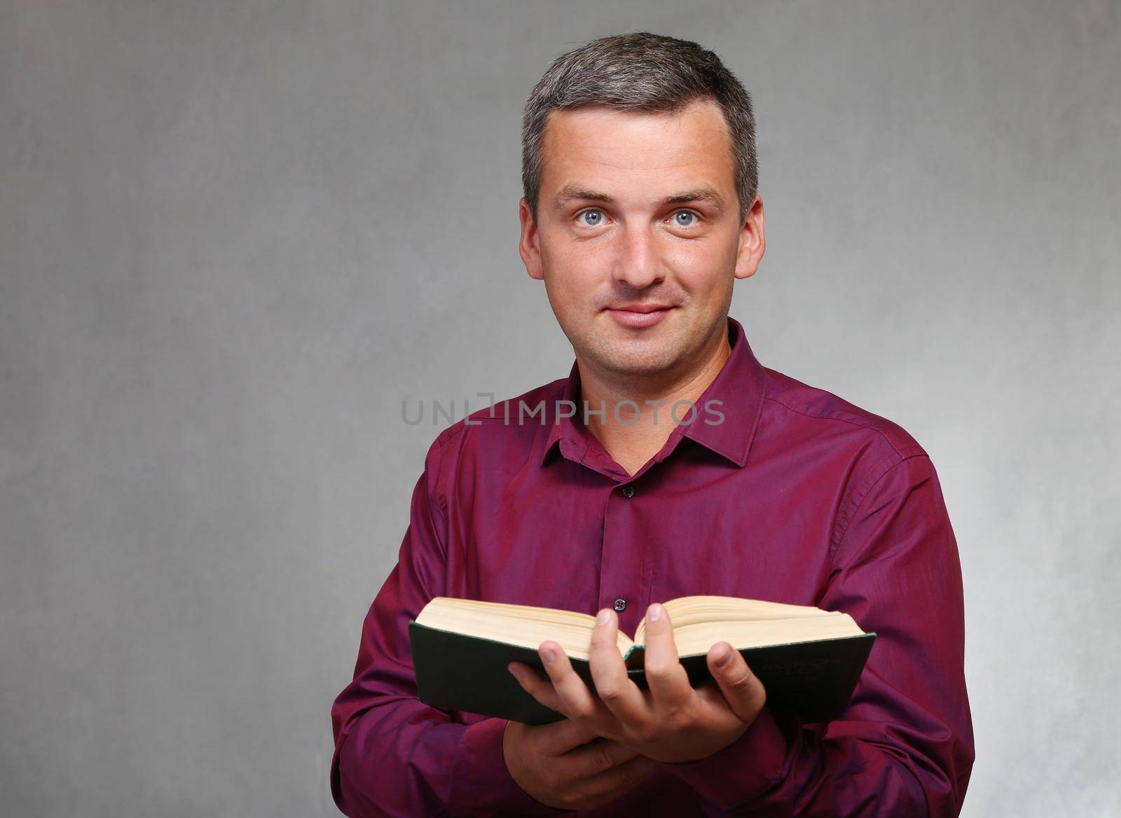 An attractive, cheerful man in a red shirt smiles and holds a book in his hand. Teacher with a book on a gray studio background. looks into the camera.