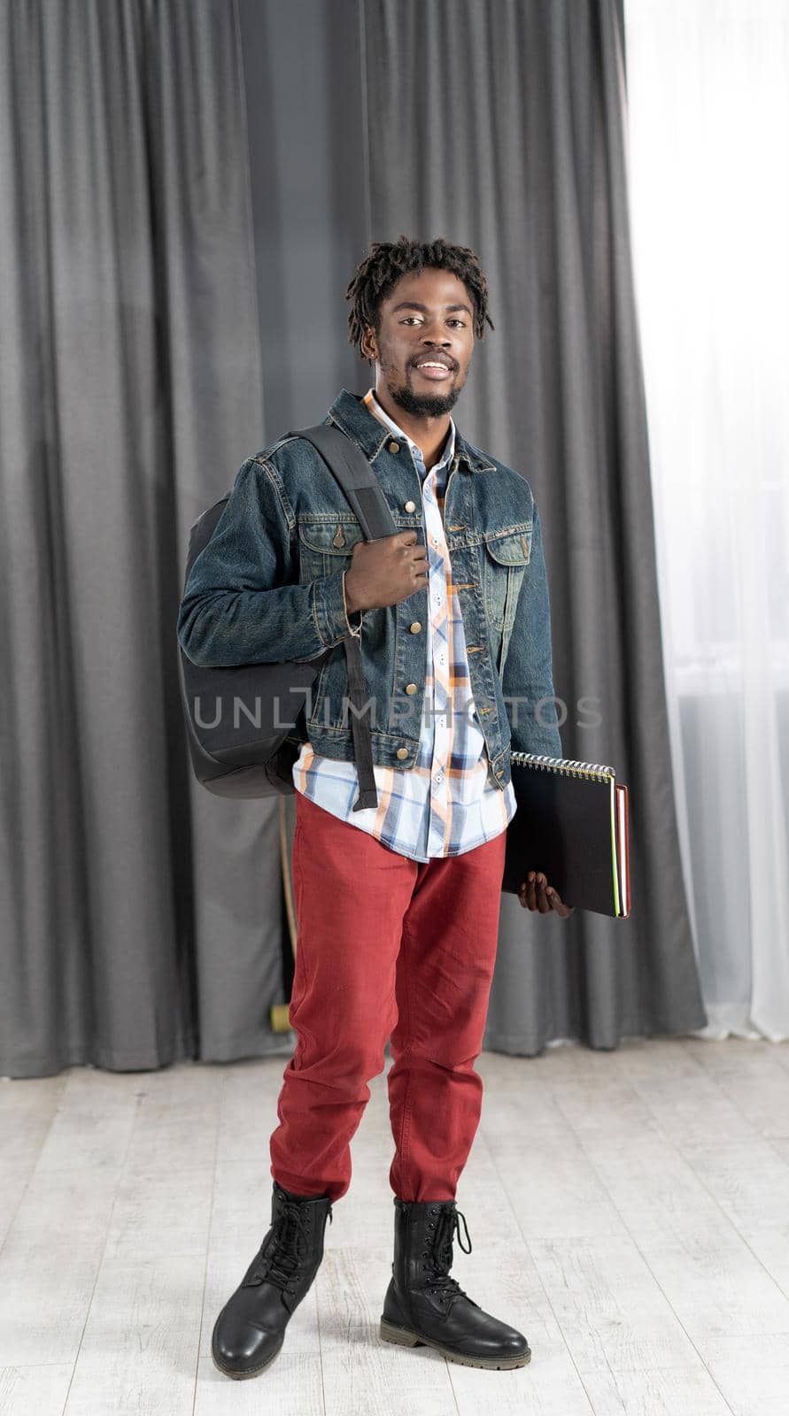 Trendy young african-american man with beard holding books and backpack wearing denim jacket ready to go to studies. Handsome young student man on the way to school. School concept. Studies concept.