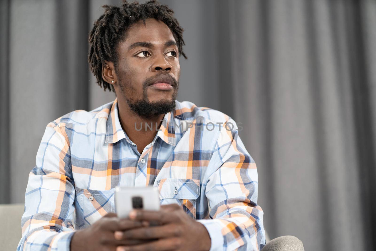 Thoughtful African American man use smartphone checking social media or updating information. Portrait of young man holding smartphone sitting on the sofa. Social media concept.