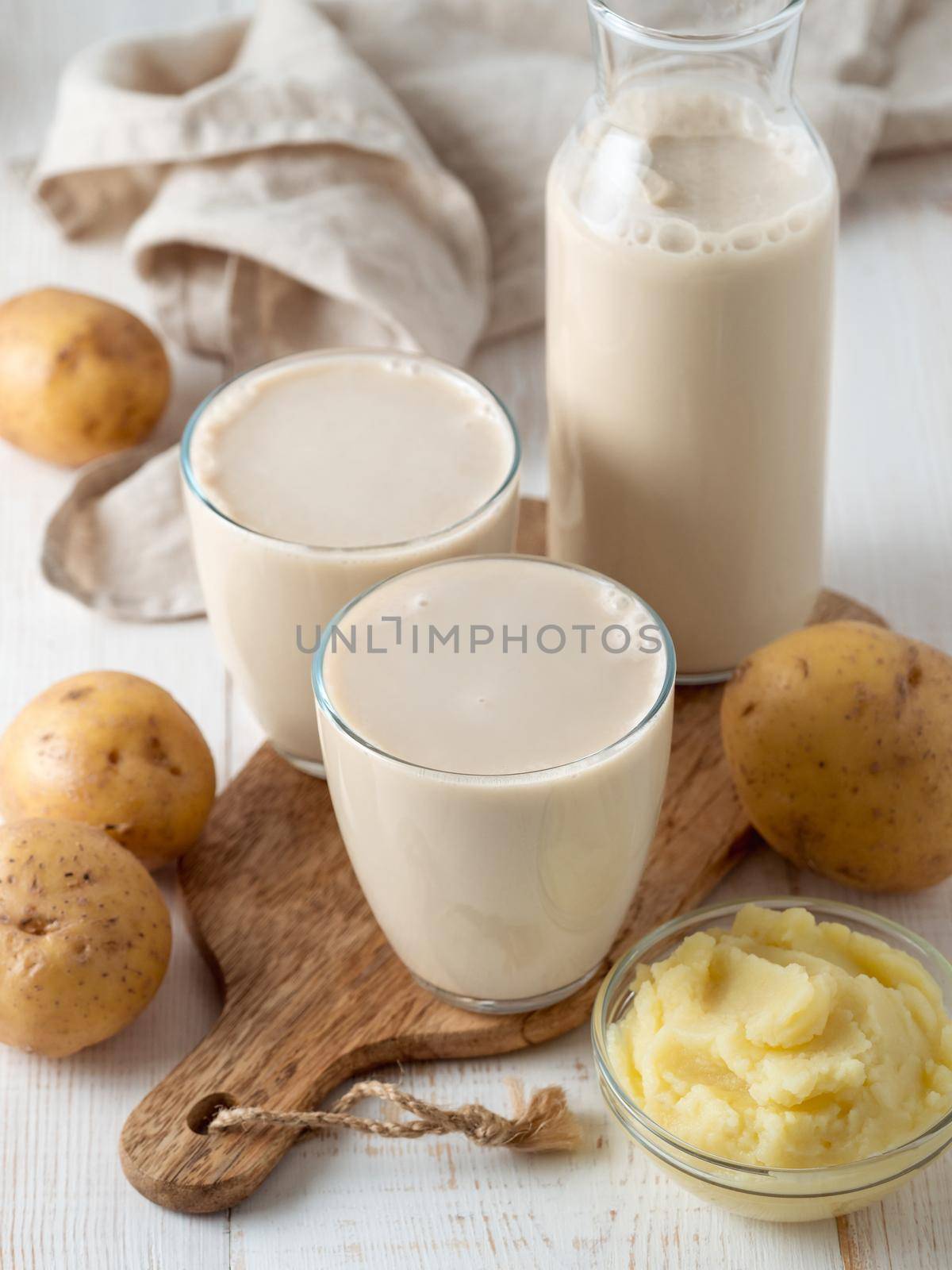 Potato milk in glasses on white wooden background. Pouring vegan milk in glass, with potato puree and potato tubers on background. Copy space. Home made potato milk made from boiled potatoes