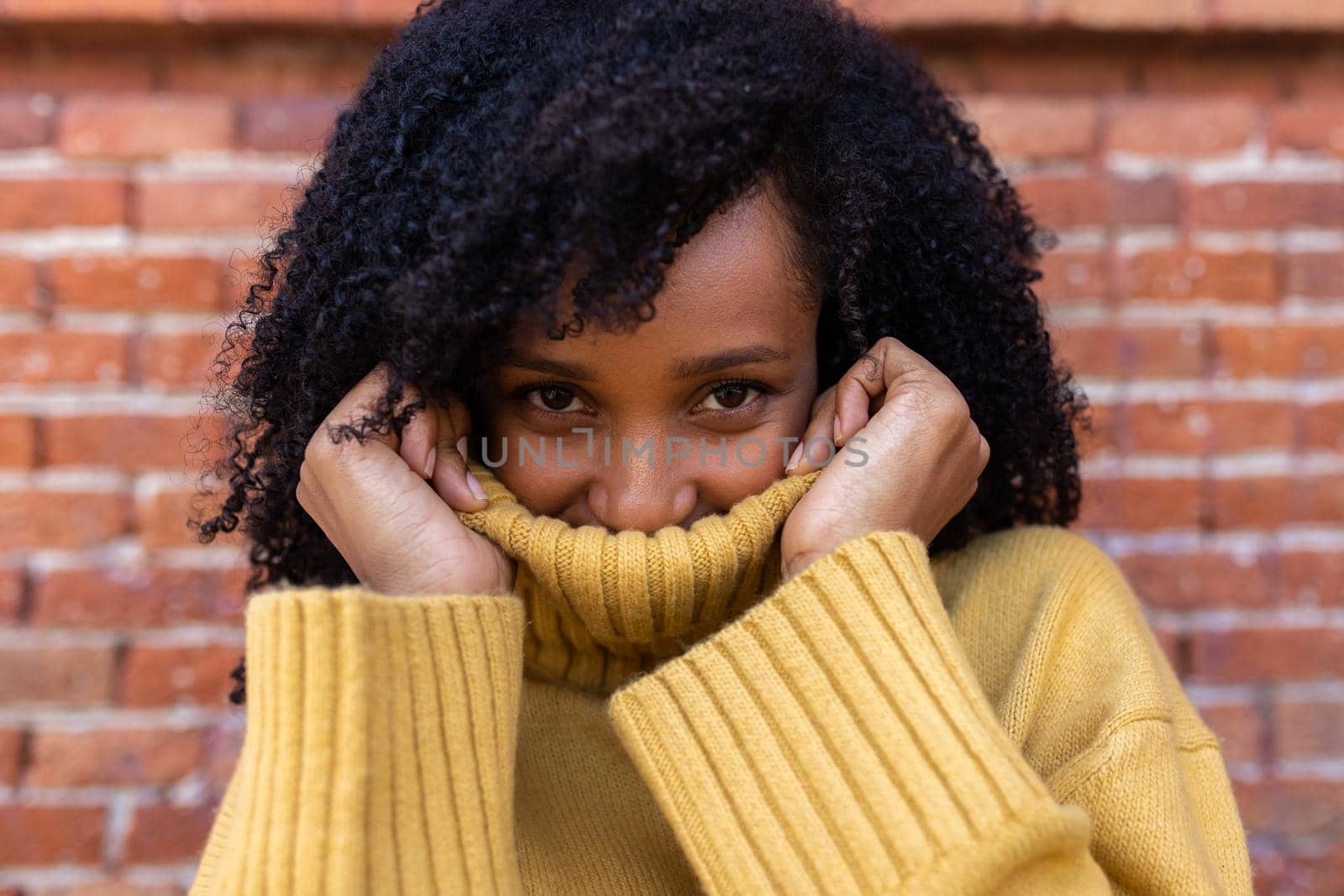 Young African American woman looking at camera covers her face with the collar of her sweater. Brick wall background. by Hoverstock