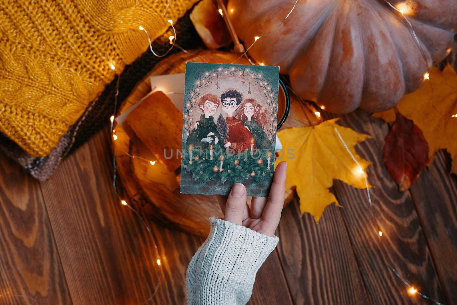 Postcard with illustration from Harry Potter in hand. Pumpkin, knitted sweater, autumn leaves and lights of garlands on a wooden background. Golden trio. Bishkek, Kyrgyzstan - November 1, 2019.