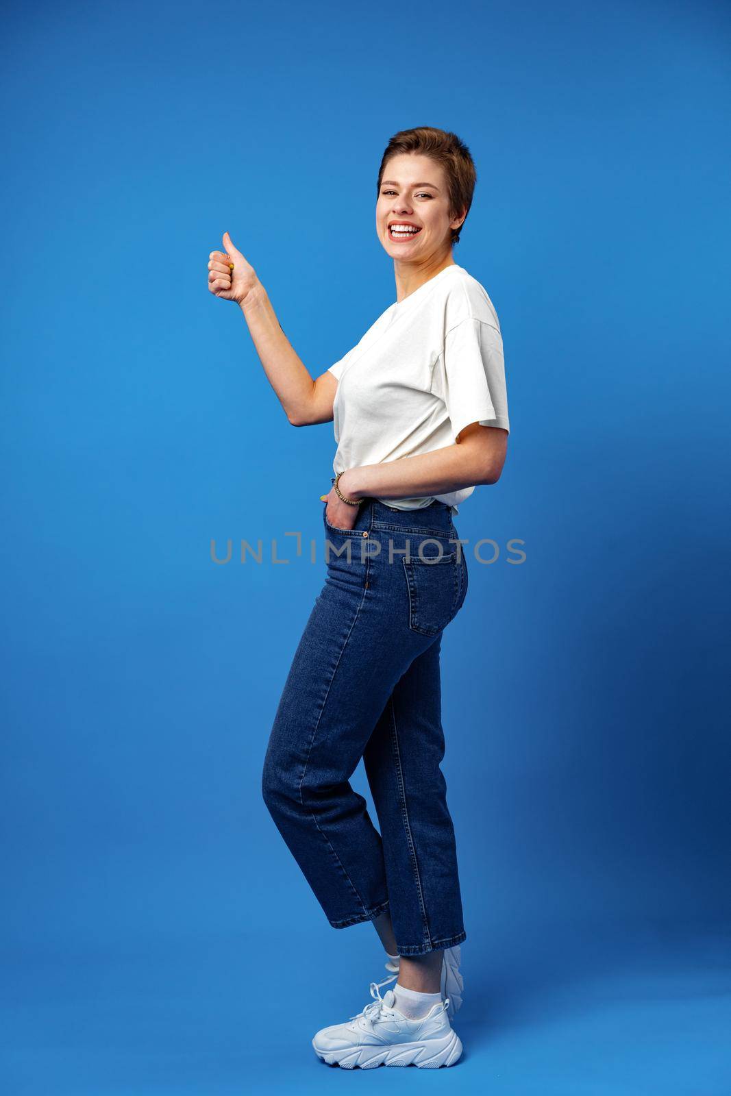 Young happy cheerful woman with short hair showing thumb up on blue background, full length portrait