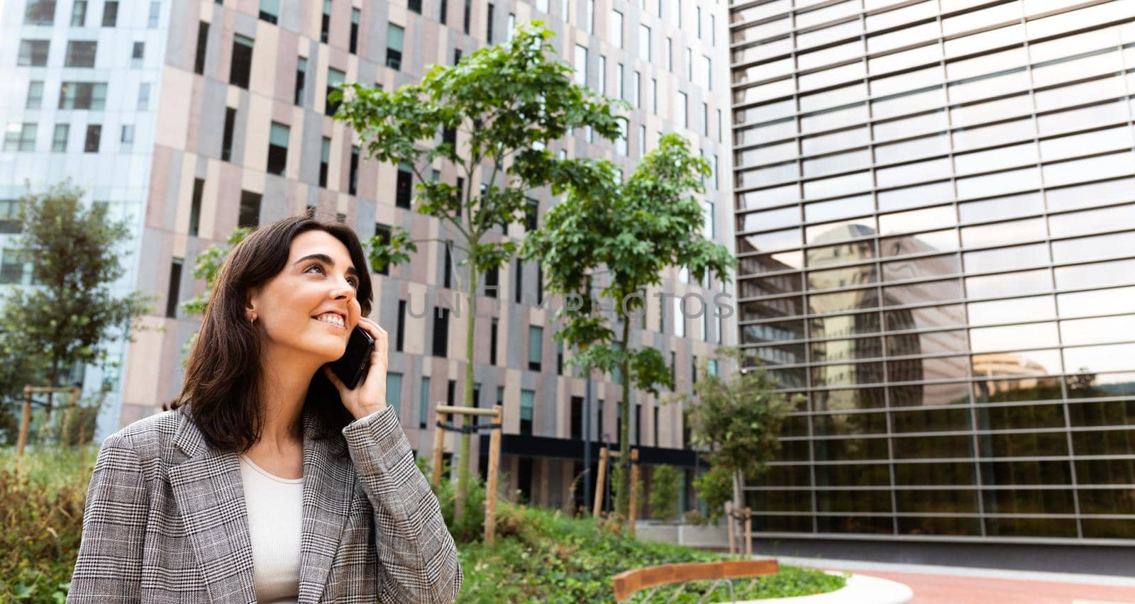 Panoramic image of smiling young business woman talking on cellphone next to office buildings. Copy space. Business and technology concept.