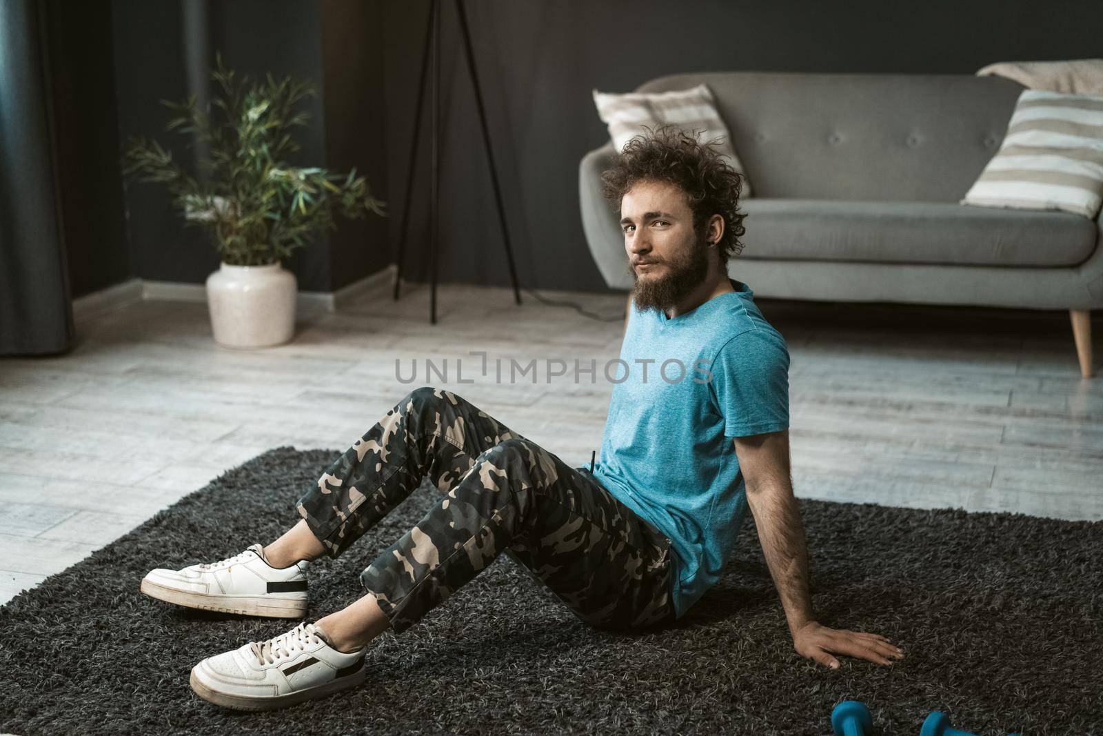 Handsome young man doing home work out sitting on the carpet relaxed at home looking at camera. Funny curly hair man with a mess beard doing exercise at home. Concept of healthy life.