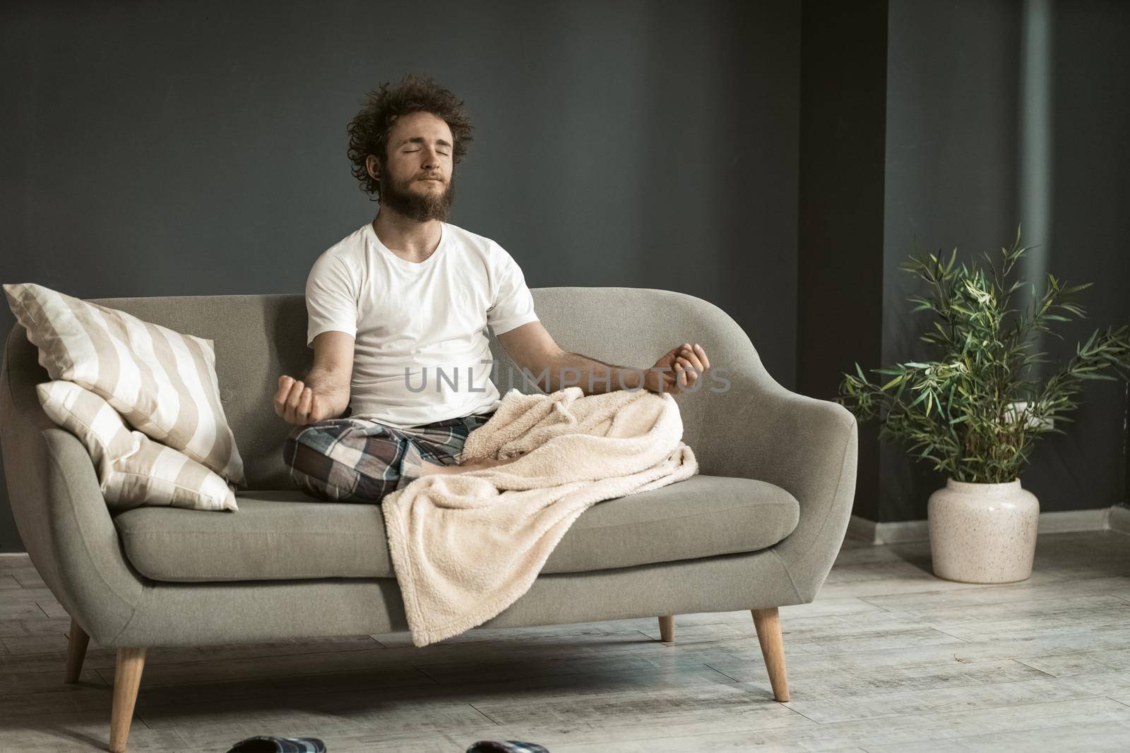 Young handsome man sitting on the couch or sofa at home enjoying meditation, relaxation, calmness, in his pajamas in living room during lockdown or being sick.