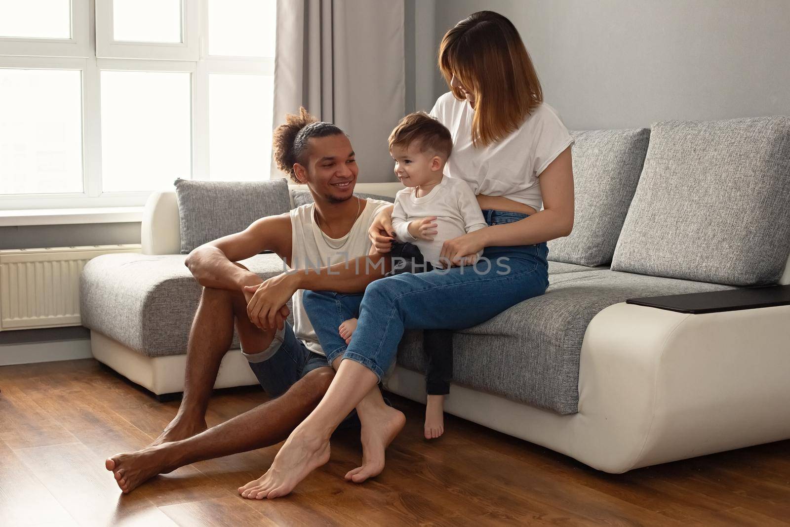 Happy, modern, multiracial family with a little boy in cozy home clothes sit on a gray sofa in romm, near a large window, smiling. Copy space