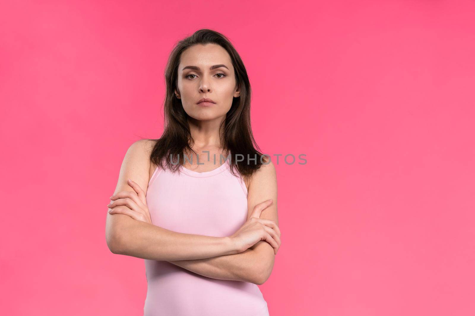 Suspicious young woman in a pink sleeveless t-shirt folded her arms and looks at the camera. Doubt, mistrust young beautiful woman isolated on pink background.