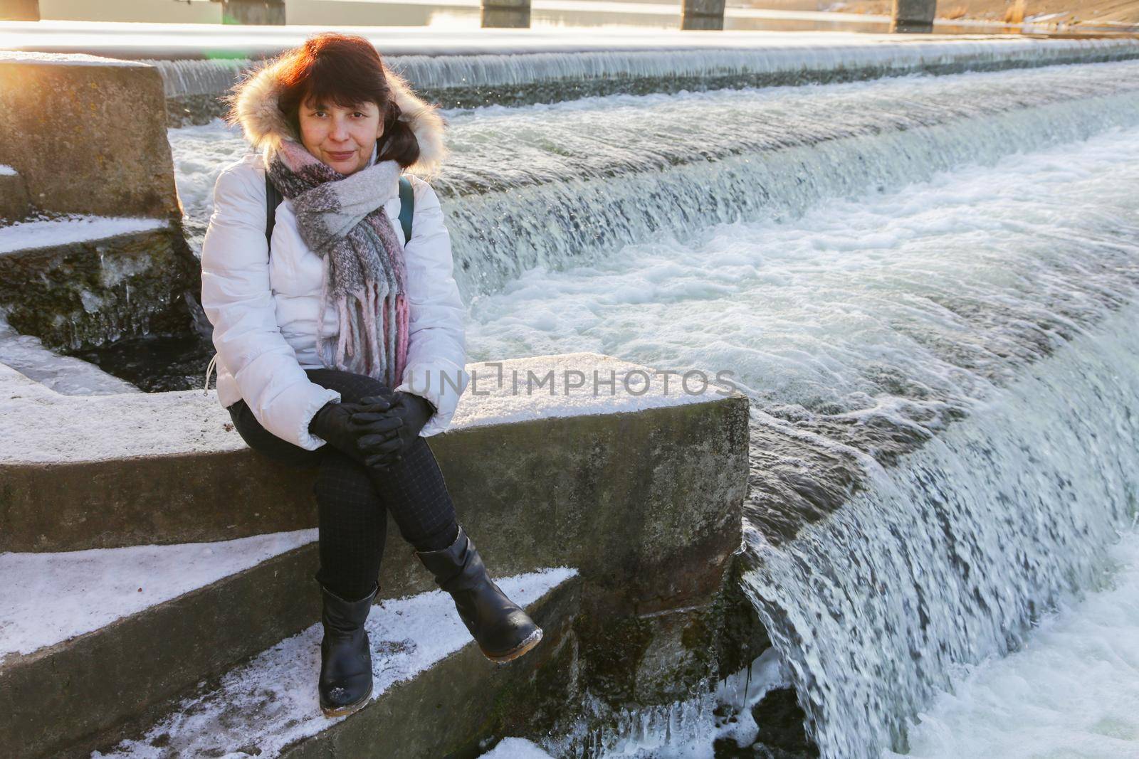 Pretty woman in a white jacket with a scarf and black gloves sitting near a waterfall at the reservoir.