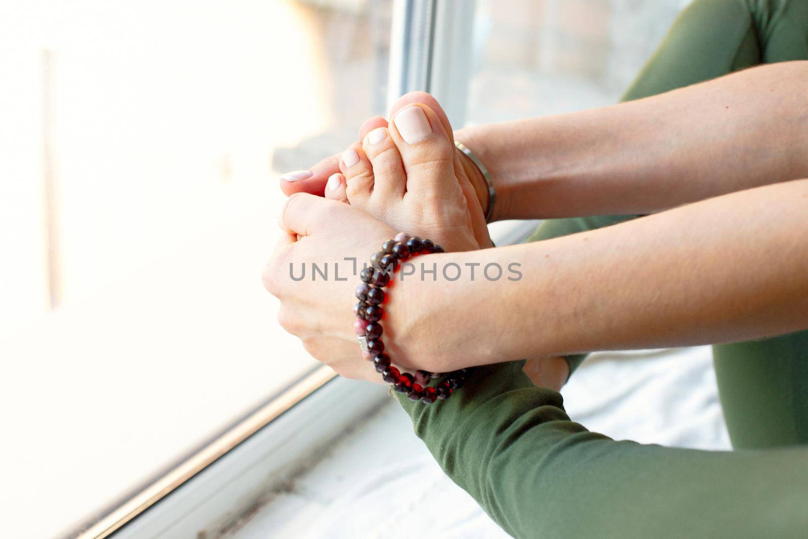A slender woman in a green jumpsuit, in a yoga pose, clasped her feet in her arms, sits by a large window. Close-up